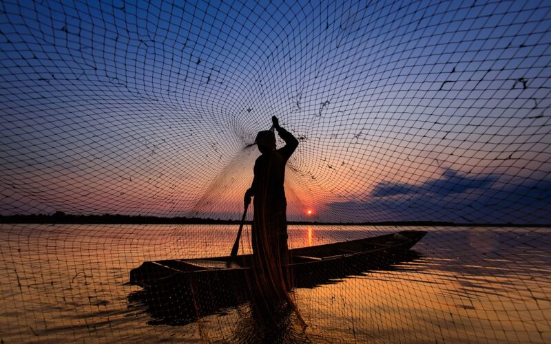 Man on a boat at dusk casting a fishing net